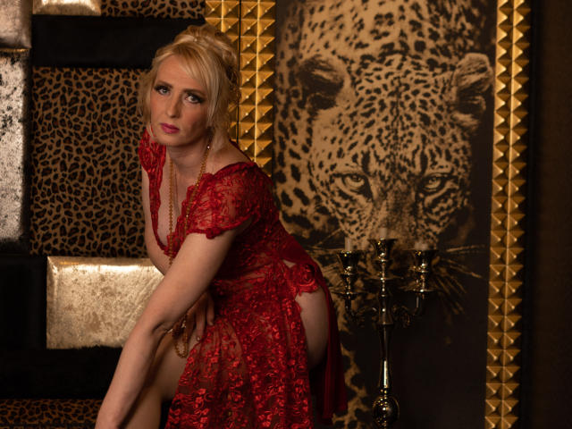 Blonde EroLayer in einem roten Spitzenkleid, das ihren Hintern zeigt, mit einer Halskette, die in die Kamera schaut, in einer Umgebung mit Leopardenmuster und Goldtönen, stehend neben einem Messing-Kerzenständer..