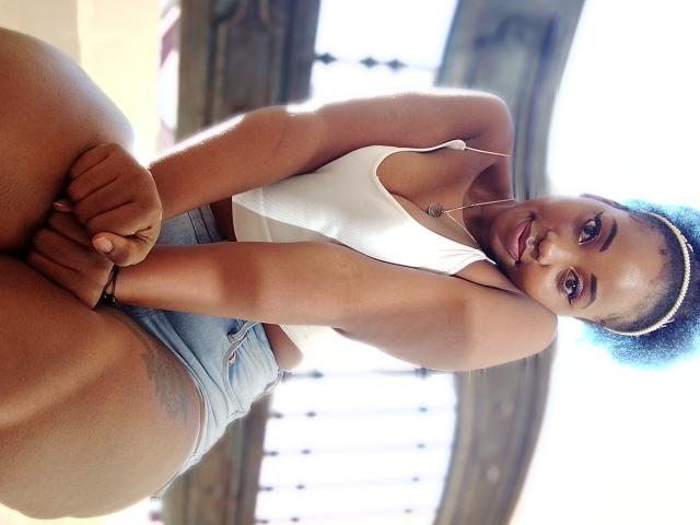 CindyQ, com pele escura, cabelo cacheado azul, camiseta branca e shorts azul claro, sentada com os braços cruzados, mostrando uma tatuagem na coxa direita, sorrindo para a câmera..