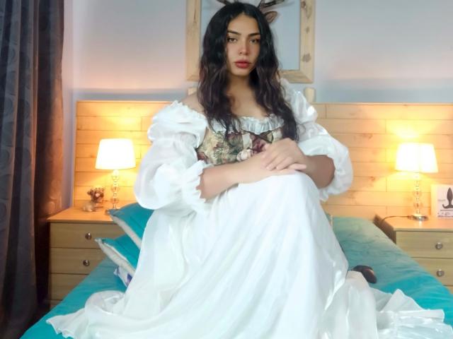 LiliithBuckland, dark-haired, in white puffy dress, sitting on bed, legs together, arms crossed, holding dress, looking at camera, wooden headboard, blue bedspread, two lit lamps.