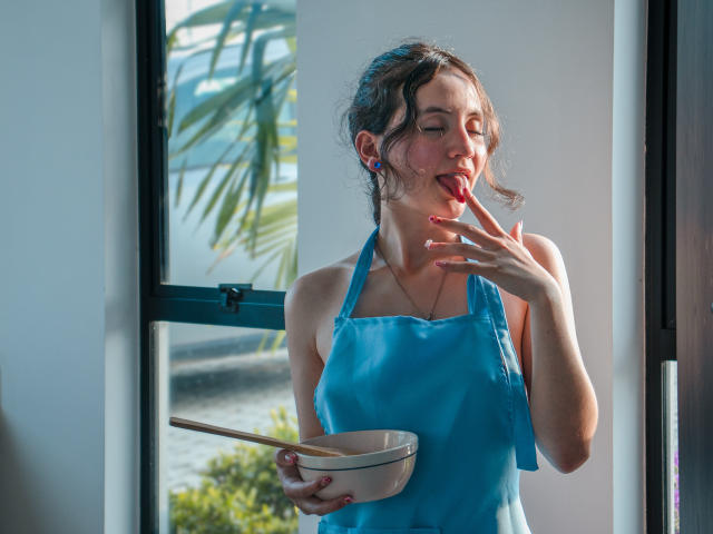 ChloeDulce, con i seni visibili sotto un grembiule blu, mangia zuppa con le bacchette, con la lingua fuori, tenendo una ciotola nella mano destra, in piedi di fronte a una finestra..