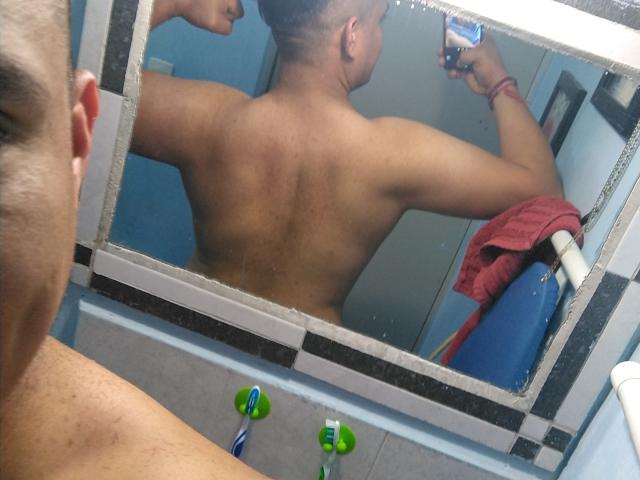 AbednegoDadreny, shirtless, with a toned back, holding a toothbrush, reflecting in a mirror. Red towel on the right, blue and green toothbrushes on the sink.