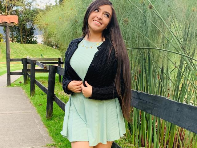 EvaMcAllister with long brown hair, smiling, wearing a green dress, black jacket, and gold necklace, standing by a black fence, grass, and tall green plants. Boobs visible.