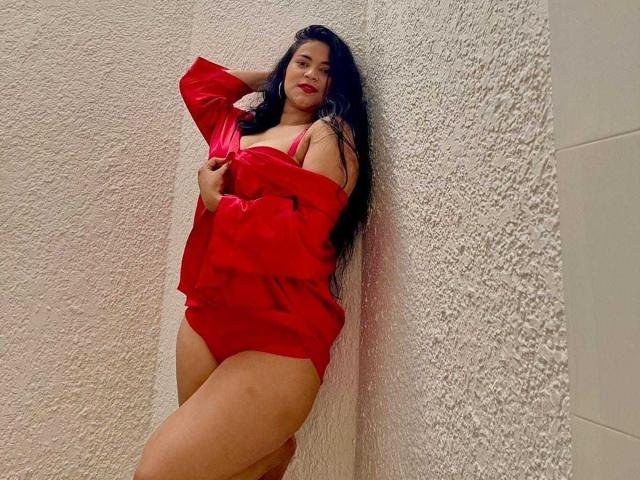 HeilinSquirt with long black hair, red shirt, and red lingerie, leaning against textured white wall, showing her curvy ass and large tits, with a confident, sexy expression.