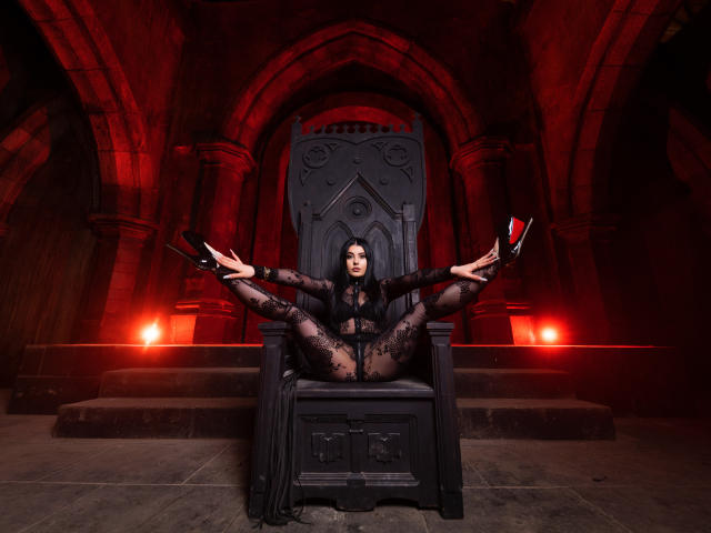 MelissaGlow, black lace bodysuit, spread legs on gothic throne, wearing red-soled black heels, dark wavy hair, red-lit medieval-style room, stone floor, seductive, confident, sexy.
