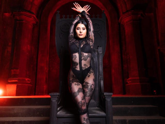 MelissaGlow with dark black lace bodysuit, showing her tits and ass, arms raised, standing in front of a large black throne, red lighting, and Gothic arches in background.