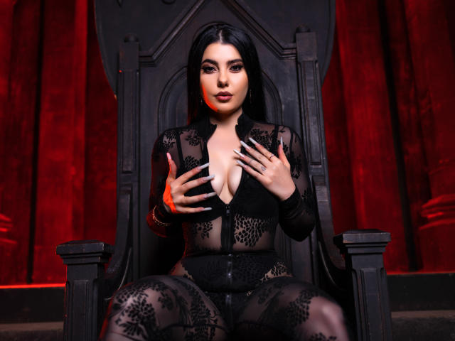 MelissaGlow, with black lace bodysuit, sits on a dark throne, hands on her tits, long black hair, red curtains, and a seductive expression. Her nails are long and painted.