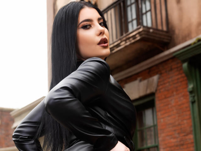 MelissaGlow with long black hair, wearing a black leather jacket, looking over her shoulder with her mouth slightly open, showing her lips and subtle makeup. Her ass is visible.