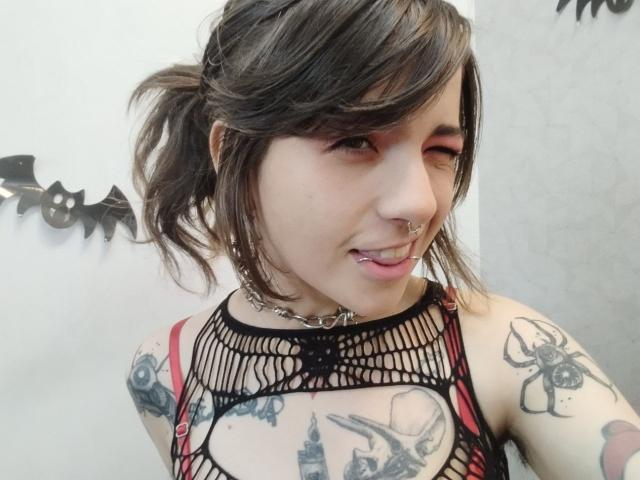 AmySpider with dark hair in a ponytail, red eyeliner, nose ring, and lip piercing, wearing a black fishnet top, showing tattoos on arms, and a choker.