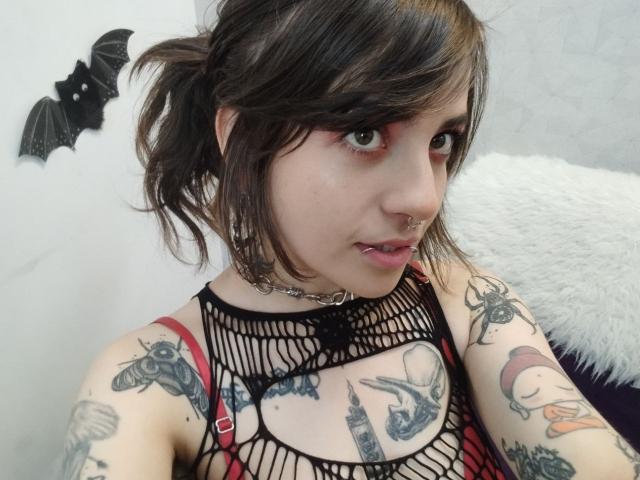 AmySpider has dark brown hair, green eyes, and multiple tattoos. She's wearing a black fishnet top, red straps, and a chain necklace. Her nipples are visible. Bat wall decoration.