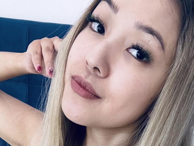 Het gezicht van RenyLime, met blond haar, donkere ogen en rode lippenstift, haar hand raakt haar hoofd, ze kijkt opzij, draagt lange wimpers, haar nagels zijn rood gelakt..