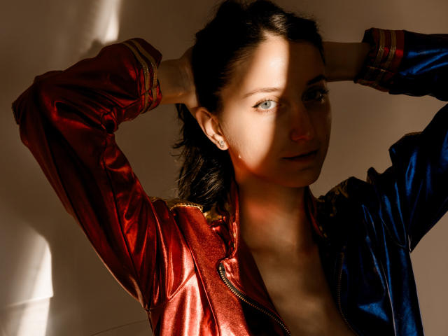 KettyBelly con las manos en el cabello negro, vistiendo una chaqueta roja y azul brillante, con la luz del sol en el rostro, una pequeña gota de sudor en la mejilla, ojos azules, una ligera sonrisa, sexy, una sombra sutil..