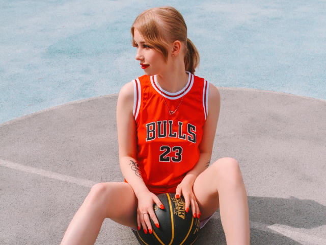 NicoleBrian con una camiseta de baloncesto roja, número 23, sentada en la cancha con las piernas separadas, sosteniendo una pelota, uñas rojas, tatuaje en el antebrazo izquierdo, coleta rubia, mirando hacia un lado..