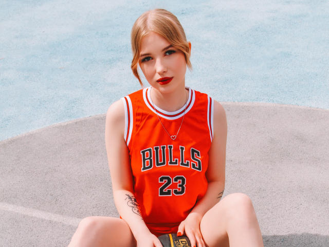 NicoleBrian, blonde, rote Lippen, sitzt auf Beton, trägt ein orangefarbenes Basketballtrikot mit "BULLS 23", hat Tattoos an Armen und Beinen, Beine gespreizt, blickt in die Kamera, trägt eine Halskette, sonniger Tag, süß..
