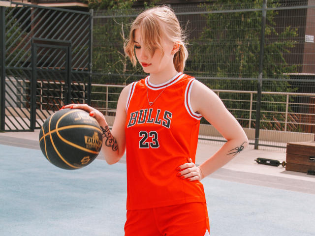 NicoleBrian, com cabelo loiro, vestindo uma camisa e shorts de basquete laranja, segurando uma bola de basquete, mostrando tatuagens no braço e na mão, em pé em uma quadra ensolarada..