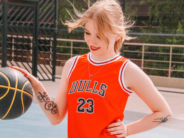 NicoleBrian with blonde hair, red lipstick, and orange Bulls basketball jersey, holding a ball, has tattoos on her right arm and left wrist, smiling on a sunny court.