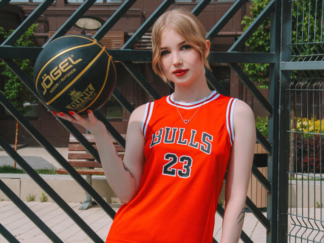 NicoleBrian with blonde hair, red lipstick, wearing an orange Bulls basketball jersey (23), holding a black Peel basketball, standing in front of a metal fence.
