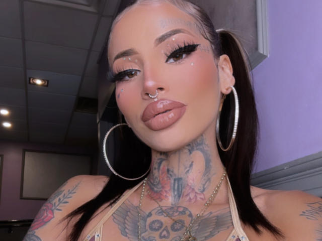 LovelyWillow, con tatuajes, piercings, grandes aros, labios brillantes, pestañas largas y coletas, mostrando sus tatuajes en el cuello y el pecho, y un piercing en la nariz..