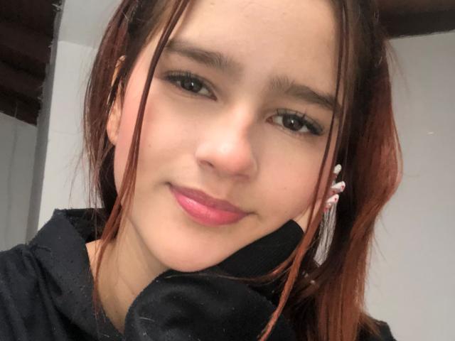MelodyMansson's face, brown eyes, pink lips, light skin, small nose, black hoodie, red-brown hair, slight smile, white ear clip, soft light, casual selfie.