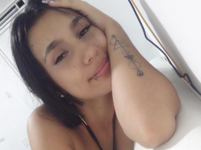 KataleyaMansson with black hair, brown eyes, and tattoos on her arm, smiling with a nose ring and lip piercing, resting her head on her hand.