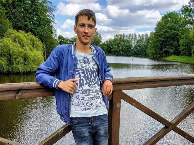 StormBernard with short brown hair, blue jacket, white shirt, and jeans, leaning on a wooden bridge, standing by a calm, green-leafed pond, under a cloudy sky.