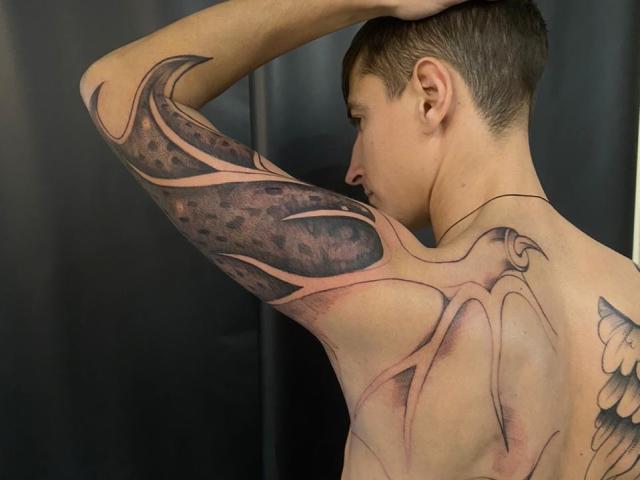 StormBernard with a large, intricate tattoo covering his right arm and upper back, showing dark, leaf-like patterns with pink lines. He's shirtless, head turned, short brown hair.