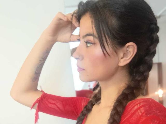 Sammy Baker, com cabelo castanho trançado, blusa de malha vermelha, braço levantado, perfil lateral, sombra rosa nos olhos, tatuagem no antebraço, olhando para a direita, lábios rosados suaves, parede branca ao fundo..
