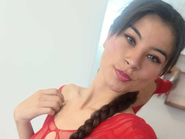 Sammy Baker, com uma blusa vermelha de malha, cabelo castanho trançado, piercing no nariz, batom rosa e um sorriso discreto, segurando a blusa, mostrando os seios. Luz suave, selfie casual..
