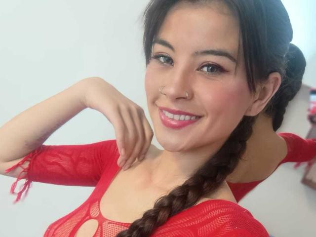 SammyBaker with a smile, red mesh top, nose ring, and braided brown hair, pink lipstick, and a cute face, showing off her tits and ass in the photo.