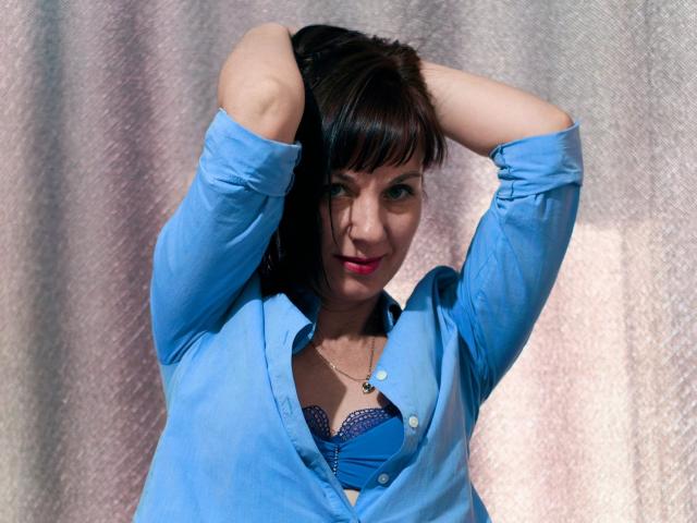 DianaAhTender, con le braccia alzate, indossa una camicia blu con bottoni, che lascia intravedere il suo reggiseno blu, ha capelli castano scuro, rossetto rosa e un sorriso appena accennato, su uno sfondo con una texture particolare..