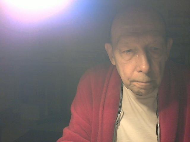 BerryX, bald with a puffy face, wearing a red zip-up and white shirt, looks tired and annoyed with a small, droopy mustache, under harsh, blinding light.
