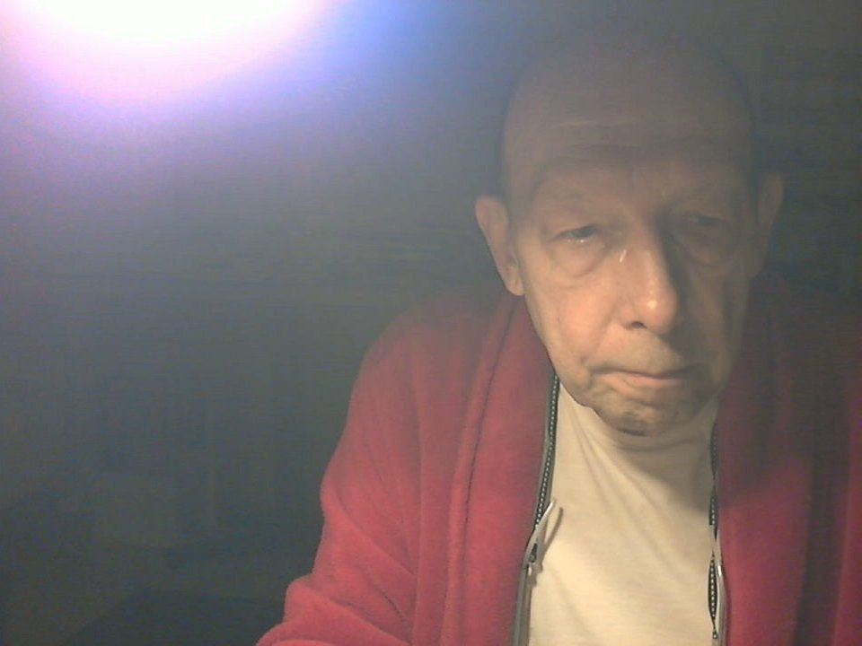 BerryX, bald with a puffy face, wearing a red zip-up and white shirt, looks tired and annoyed with a small, droopy mustache, under harsh, blinding light.