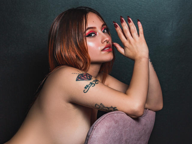 Antokira with reddish-brown hair, red eyeshadow, and dark nail polish, topless, showing tattoos on her arm, leaning on a purple chair, hands near face.