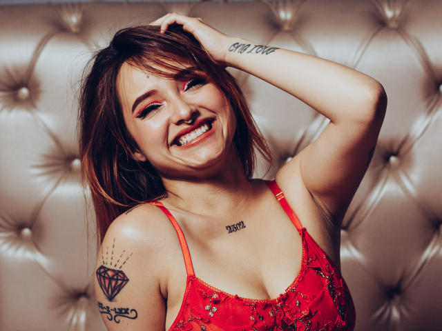 Antokira with smiling face, red lace top, brown hair, tattoos on shoulder and chest, hand in hair, visible tits, pink eyeshadow, tan leather background, cute, sexy.