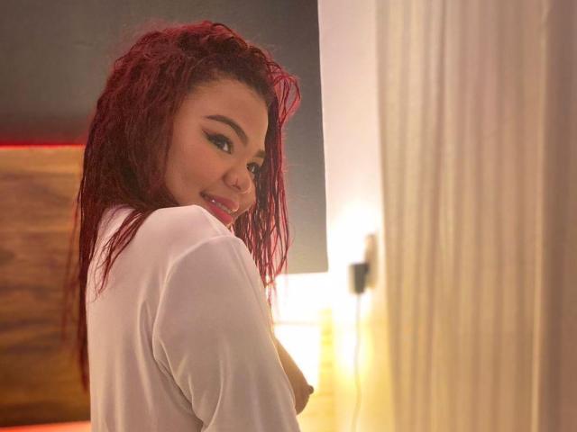 MiahEvaanss with red curly hair, white shirt, looking over shoulder, smiling, showing a small part of her tits, in a softly lit room, with a warm light.