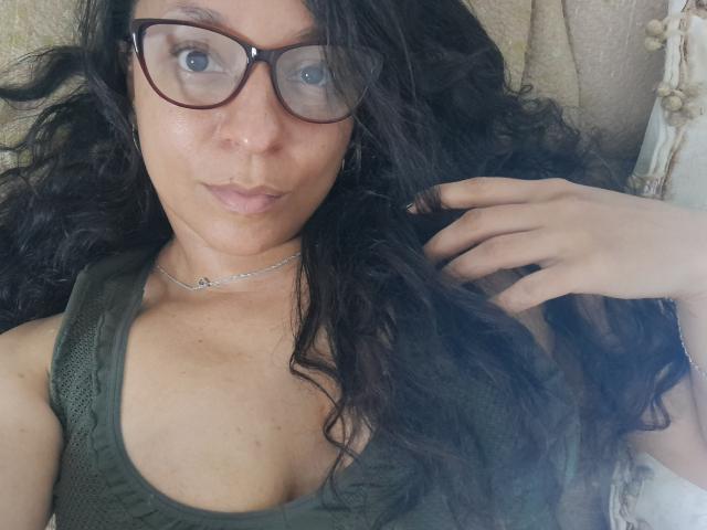 Angelonix a les cheveux noirs et bouclés, elle porte des lunettes, un débardeur vert et un collier en argent. Elle est allongée, une main dans les cheveux, et regarde l'appareil photo..