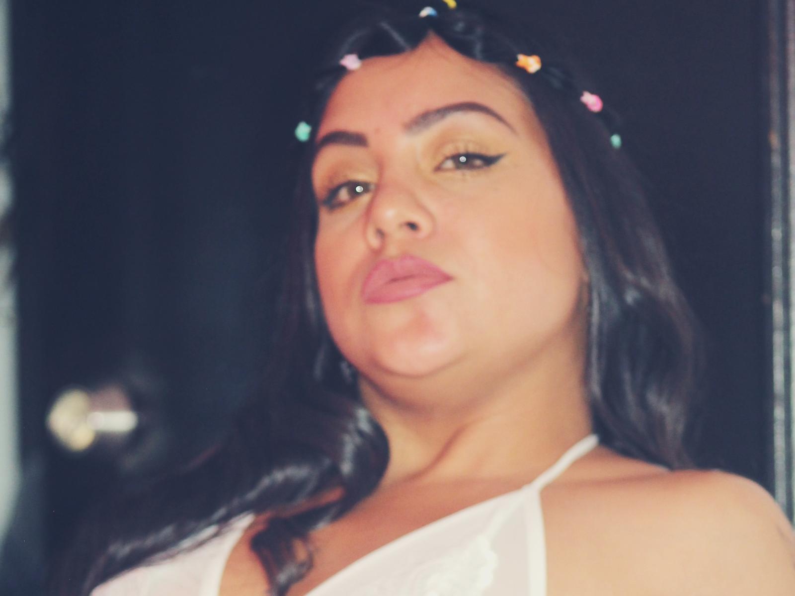 EvaCandy69, met donker haar, draagt een witte top met halter en heeft kleurrijke kerstverlichting in haar haar, volle, roze lippen en eyeliner met een vleugje, en ze kijkt er een beetje verveeld uitzien..