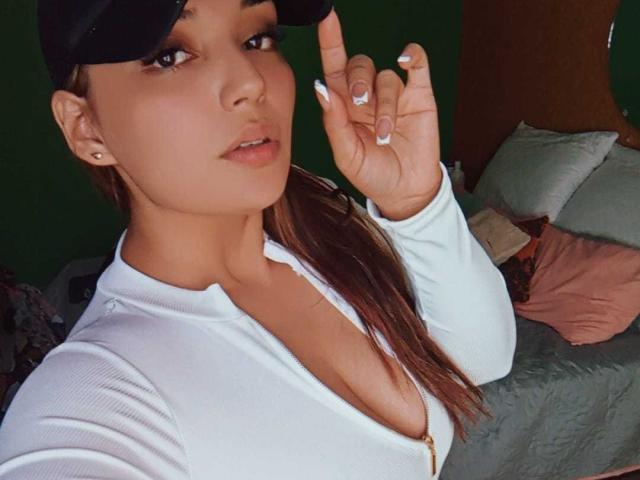 AbigaillMorrys with tan skin, brown hair in a ponytail, wearing a black cap, white zip-up, showing her cleavage, holding up her finger, with a sexy, confident look.