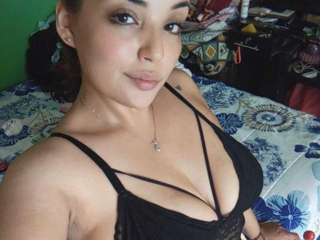 AbigaillMorrys with tan skin, dark hair in a bun, wearing a black lace top, showing her big tits, necklace, and a subtle smile, sitting on a floral bed.