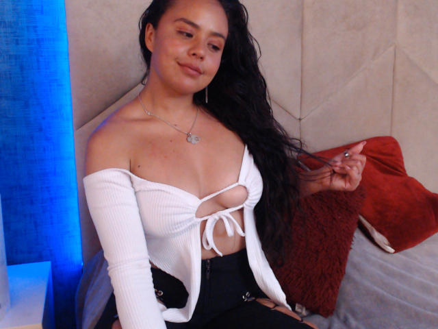 Arielle Wilde, con lunghi capelli neri, indossa una maglietta bianca a spalle scoperte che rivela il seno, pantaloni neri, seduta su un letto, con un cuscino rosso, illuminazione blu, una collana d'argento, sorridente, mentre si tiene i capelli..