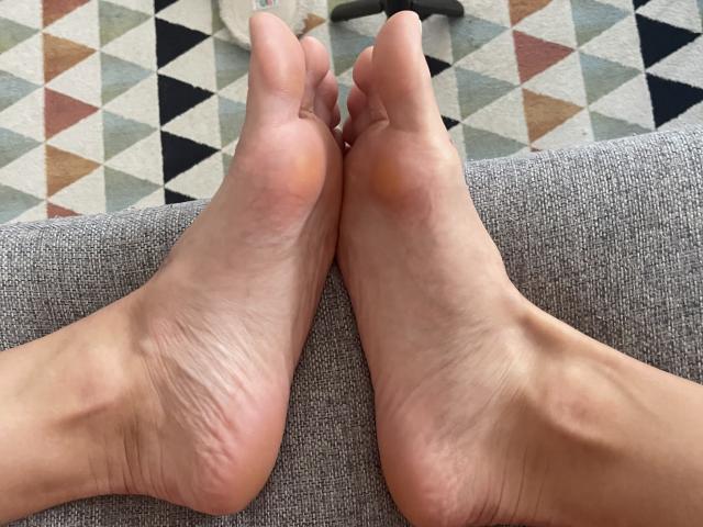 Le piante dei piedi, i piedi e le dita di SandraSwweet sono appoggiati su un divano grigio con una texture particolare, con una pelle leggermente segnata da rughe e con le linee del tallone ben visibili..