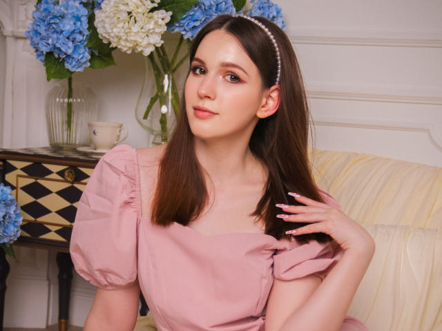 JuliaKing with brown hair, pink dress, puffed sleeves, pearl headband, subtle makeup, and pink nails, sitting on a cream sofa, hand on chest, blue and white hydrangeas in background.