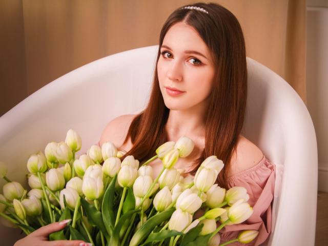 JuliaKing with straight brown hair, pink off-shoulder top, holding a bunch of white tulips in a white chair, smiling softly. She's got cute tits and a delicate face.