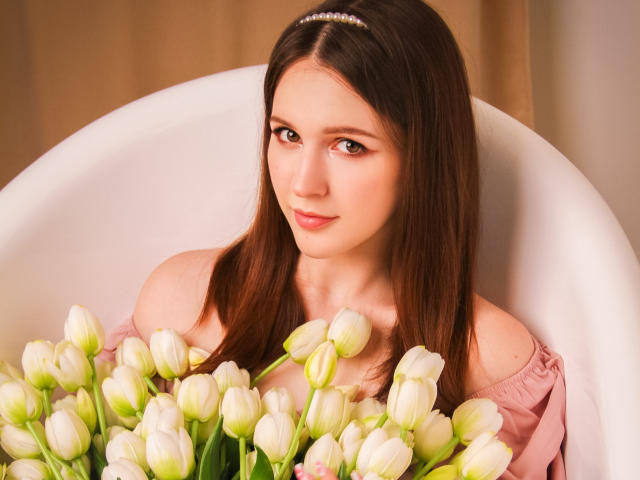 JuliaKing with brown hair, wearing a pink off-shoulder top, sitting in a white chair, holding a bunch of white tulips, looking up with a gentle smile.