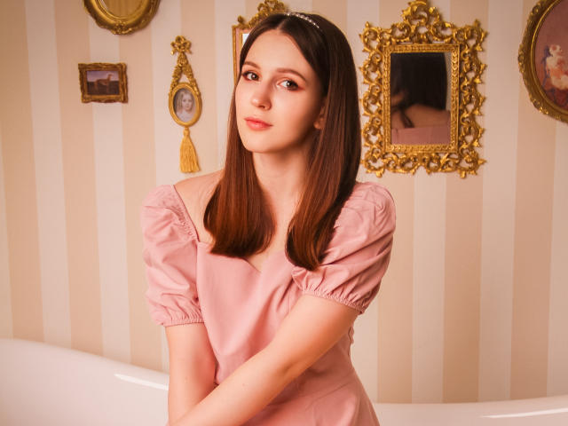JuliaKing with straight brown hair, wearing a pink puffed-sleeve dress, standing in a room with gold-framed mirrors and striped wallpaper, arms crossed, looking slightly to the side.