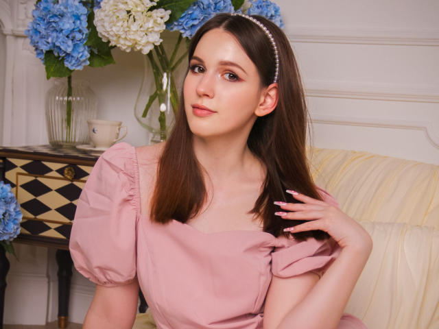 JuliaKing with straight brown hair, pink dress, puffed sleeves, pearl headband, and pink nails, sitting on a cream couch, looking slightly up with a soft smile.