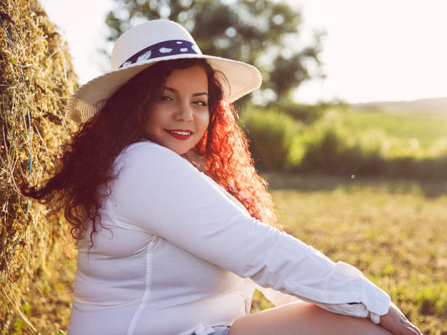 MiryamInes, con cabello rizado y pelirrojo, vestida con una camisa blanca de manga larga y un sombrero de sol con una banda azul, sentada contra un fardo de heno, sonriendo y con lápiz labial rojo..