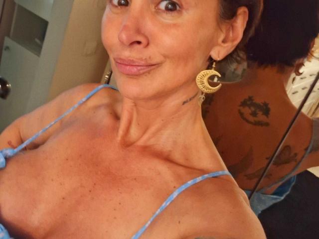 BriliantOne with tan skin, small tits, and a slight smile, wearing blue straps, has gold earrings, visible neck tattoo, and reflection showing a large back tattoo.
