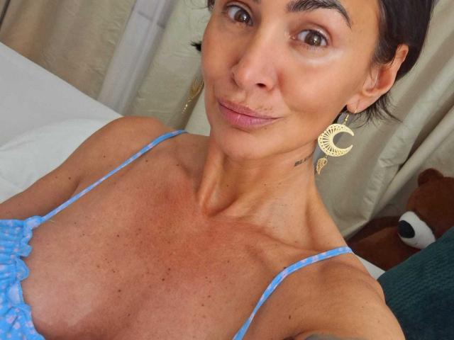 BriliantOne with tan skin, brown eyes, and small tits wearing a blue polka-dot top and crescent moon earrings, smiling slightly. Brown hair, subtle makeup, and a tattoo on neck.
