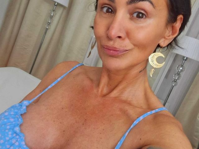 BriliantOne has tan skin, brown eyes, and dark hair, wearing a blue polka-dot top, showing small tits, with gold crescent earrings, smiling slightly.