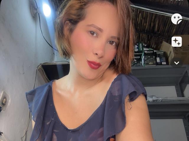 Sarita Velez mit kurzen, braunen Haaren, rosiger Rouge, rotem Lippenstift und einem blauen, gerafften Oberteil, die in die Kamera schaut und leicht die Lippen zusammenpresst. Ihre Brüste sind klein, und sie ist süß..